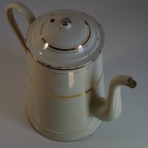 Könnte beinhalten: Eine weiße Emaille-Kaffeekanne mit goldener Verzierung. Die Kanne hat einen Deckel und einen Ausguss.
