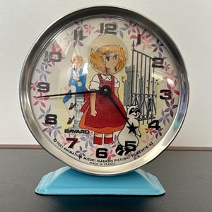 Può includere: Un orologio da tavolo argentato con un'immagine a cartone animato colorata di una ragazza e un procione. Il quadrante dell'orologio ha uno sfondo bianco con numeri neri e una lancetta delle ore e dei minuti nere. L'orologio ha una base blu e il testo "OTOEI ANIMATION MIZUKI IGARASHI PICTURAL FILMS 1978 A1 MADE IN FRANCE" è stampato sul fondo.