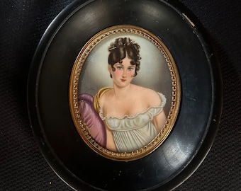 May include: A framed miniature portrait of a woman with dark hair, wearing a white dress with a purple shawl. The portrait is set in a black oval frame with a gold trim.