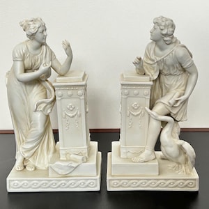 May include: Two white porcelain statues of figures standing on pedestals. The figure on the left is a woman with her arm raised, the figure on the right is a woman with a swan at her feet.