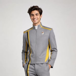 Op de afbeelding: Een grijs uniformset bestaande uit een jack met ritssluiting en opstaande kraag en een bijpassende broek. Het jack is voorzien van gele en zwarte verticale strepen, een witte binnenkraag en een metalen delta-vormig embleem op de linkerborst.