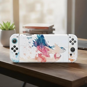 Puede incluir: Consola Nintendo Switch blanca con una funda personalizada con un diseño de acuarela de una criatura mítica en tonos azules, rosas y morados. El diseño incluye estrellas y un efecto de salpicadura. La consola está sobre una superficie de madera.
