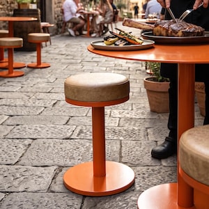 Pode incluir: Uma mesa redonda de metal laranja com um banco de assento acolchoado bege, onde uma pessoa com avental preto e o texto ETÇI corta um bife grande cozinhado num prato ao lado de um copo de vinho branco numa área de refeições ao ar livre.