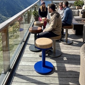 May include: A series of stools with circular, tan-coloured padded seats and vibrant blue cylindrical bases. The stools are arranged on a wooden deck, featuring a sturdy metal construction with a flat, circular floor plate for stability and support.