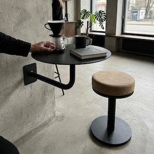 Pode incluir: Uma mesa circular preta montada na parede com um braço de suporte de metal e um banco a condizer em preto e bege. Sobre a mesa estão um coador de café de vidro, uma caneca preta e um livro fechado. O mobiliário tem um acabamento mate.