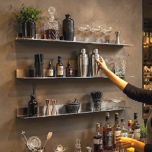 Puede incluir: Tres estantes de pared horizontales de acero inoxidable que contienen diversos utensilios de bar, como decantadores de vidrio, cocteleras de metal, vasos mezcladores, medidores y botellas, montados en una pared oscura texturizada.