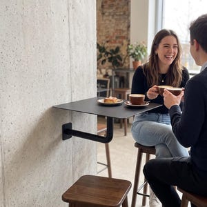 May include: A matte black wall-mounted table with a square top and a single L-shaped support bracket attached to a concrete pillar. The table holds two brown ceramic mugs filled with coffee and a small white plate with a pastry.