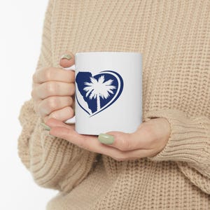 May include: White ceramic mug featuring a navy blue heart design with a palm tree silhouette. The mug is held by a person wearing a beige sweater. The design is centered on the mug.