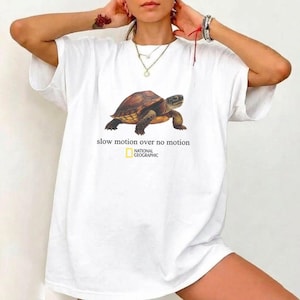 Puede incluir: Una camiseta blanca de manga corta con un gráfico de una tortuga marrón en el centro. Debajo de la tortuga, el texto dice "slow motion over no motion" seguido del logotipo de National Geographic en amarillo y negro.