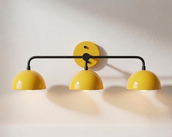 May include: A wall-mounted light fixture featuring a horizontal black metal bar attached to a circular yellow backplate. Three dome-shaped yellow shades are suspended from the bar, each housing a light bulb that emits a soft glow.