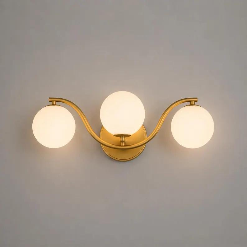 May include: A gold-toned wall sconce with three spherical, frosted glass shades. The fixture has a curved design and is mounted on a circular base. The light fixture is designed for indoor use.