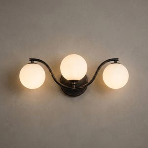 May include: A three-light vanity fixture with a dark bronze finish. The fixture features three spherical, frosted glass shades. The shades are arranged in a wave-like pattern, attached to a curved metal frame.