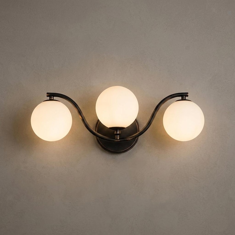 May include: A three-light vanity fixture with a dark bronze finish. The fixture features three spherical, frosted glass shades. The shades are arranged in a wave-like pattern, attached to a curved metal frame.