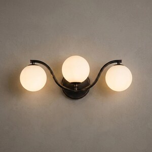 May include: A three-light vanity fixture with a dark bronze finish. The fixture features three spherical, frosted glass shades. The shades are arranged in a wave-like pattern, attached to a curved metal frame.