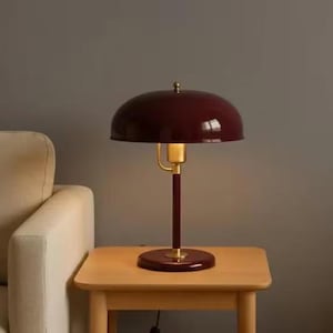 Könnte beinhalten: Eine bordeauxfarbene Schreibtischlampe mit rundem Schirm, Messingakzenten und einem dunklen Holzsockel. Die Lampe steht auf einem kleinen, hellen Beistelltisch aus Holz neben einem beigen Sofa. Das Design der Lampe erinnert an Vintage-Stile.