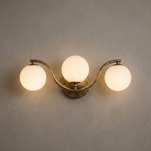 May include: A three-light wall sconce with a brass finish. The fixture features three spherical, frosted glass shades. The sconce has a curved design and is mounted on a neutral-colored wall.