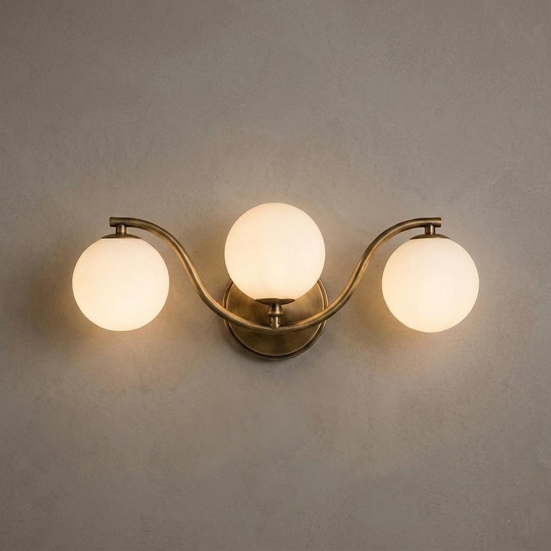 May include: A three-light wall sconce with a brass finish. The fixture features three spherical, frosted glass shades. The sconce has a curved design and is mounted on a neutral-colored wall.