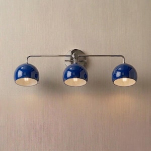 Peut inclure: Un luminaire de vanité à trois lumières avec une finition chromée polie. Il comporte trois abat-jours sphériques bleu foncé avec des intérieurs blancs. Les lumières sont montées sur une barre horizontale et une plaque arrière circulaire.