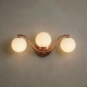 May include: A three-light wall sconce with a copper finish and three spherical, frosted glass shades. The fixture has a curved design and is mounted on a wall. The sconce provides ambient lighting.