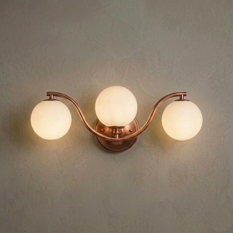 May include: A three-light wall sconce with a copper finish and three spherical, frosted glass shades. The fixture has a curved design and is mounted on a wall. The sconce provides ambient lighting.