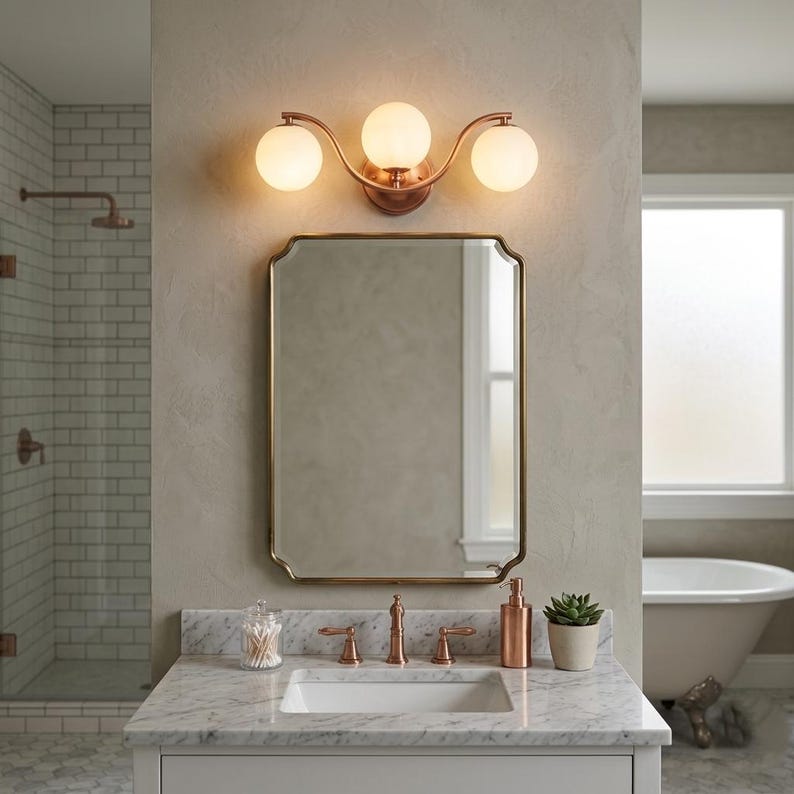 May include: A bathroom vanity with a copper-toned three-light fixture above a rectangular mirror with a gold frame. The vanity has a marble countertop with a sink, faucet, and accessories. A shower and a bathtub are visible in the background.