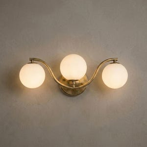 May include: A three-light wall sconce with a brass finish. The fixture features three spherical, frosted glass shades arranged in a gentle wave pattern. The sconce is mounted on a neutral-colored wall.