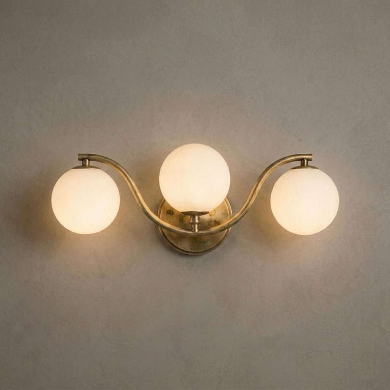 May include: A three-light wall sconce with a brass finish. The fixture features three spherical, frosted glass shades arranged in a gentle wave pattern. The sconce is mounted on a neutral-colored wall.