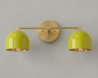 May include: A modern wall sconce with two lime green dome-shaped shades. The shades are mounted on a brass-colored arm and a circular backplate. The interior of the shades is gold, and the fixture is against a gray background.