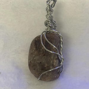 Könnte beinhalten: Handgefertigter Anhänger mit einem glatten, oval geformten Stein in Braun- und Grautönen. Der Stein ist mit silberfarbenem Draht umwickelt, wodurch ein dekorativer Käfig und eine Schlaufe zum Aufhängen entstehen. Für Halsketten.