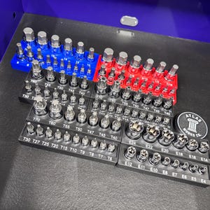 May include: A collection of various metal socket bits and torx bits, organized in black, red, and blue trays. The bits are labeled with sizes and measurements. A circular logo is visible.
