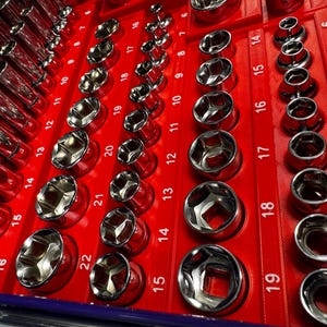 May include: A set of chrome sockets arranged in a red organizer. The sockets are various sizes, labeled with numbers, and designed for use with a wrench. The image shows a close-up view of the tools, highlighting their shiny, reflective surfaces.