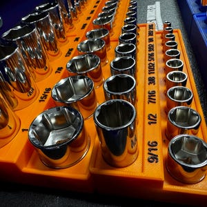 May include: A collection of polished chrome sockets neatly arranged in an orange organiser. The sockets are of different sizes, with measurements such as 9/16, 1/2, and 7/16 clearly marked. The organiser is placed inside a blue container.