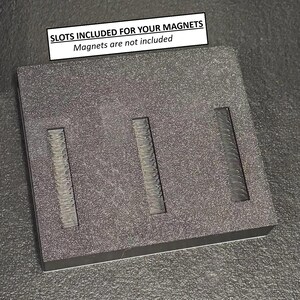 May include: A dark gray rectangular block with three rectangular slots. The block has a textured surface. A white rectangle at the top reads "SLOTS INCLUDED FOR YOUR MAGNETS" and "Magnets are not included".