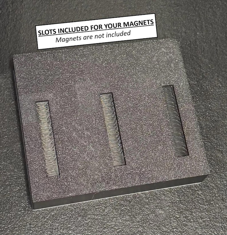 May include: A dark gray rectangular block with three rectangular slots. The block has a textured surface. A white rectangle at the top reads "SLOTS INCLUDED FOR YOUR MAGNETS" and "Magnets are not included".