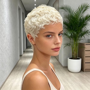 May include: A person with a short, platinum blonde pixie haircut featuring soft, wavy texture. The hair is styled close to the head with a natural, layered appearance. The individual has a neutral expression, visible shoulders, and light skin tone.
