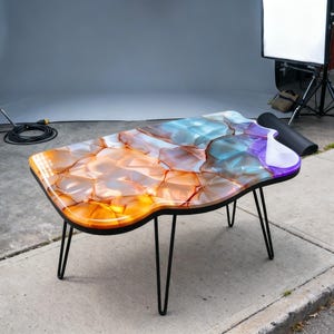 May include: A unique coffee table with a colorful, abstract design. The tabletop features a mosaic-like pattern in shades of orange, blue, and purple, with a glossy finish. It has a black border and is supported by black hairpin legs. The table is approximately 48 inches long.
