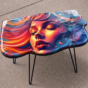 May include: A unique coffee table featuring a vibrant, artistic design. The tabletop showcases a colorful portrait of a woman with closed eyes, with flowing hair in shades of orange, red, and blue. The table has black hairpin legs.