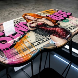 May include: A unique, artistic stool featuring a cartoon woman with red hair and a burgundy dress, set against a backdrop of a dollar bill design. The stool has a black metal frame and graffiti-style text in pink and orange.