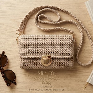 Può includere: Una borsa a tracolla all'uncinetto color beige di forma rettangolare, con una lunga tracolla abbinata e una chiusura a pressione in metallo dorato. Il testo sull'immagine recita: Mini BY crossbody bag, AMDESIGN, Skill level: advanced beginner.