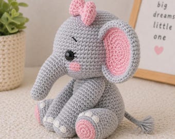 May include: A gray crocheted elephant with pink accents on the ears, feet, and cheeks, and a pink bow on its head. The elephant has black eyes and is sitting down. The background includes a framed print with the words "big dreams little one."
