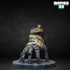 Puede incluir: Una detallada escultura de una mano de zombi verde y gris con hueso expuesto y envuelta en vendas beige. La mano descansa sobre una base redonda, texturizada, azul-gris. La imagen incluye el texto "NORAS 3D" en la esquina superior derecha.