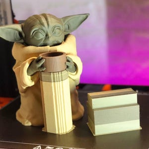 Pode incluir: Uma figura impressa em 3D de um personagem de uma popular franquia de fic&ccedil;&atilde;o cient&iacute;fica, segurando um objeto cil&iacute;ndrico. A figura &eacute; verde com orelhas grandes e est&aacute; vestindo uma t&uacute;nica marrom. Uma pilha de objetos retangulares est&aacute; ao lado da figura.
