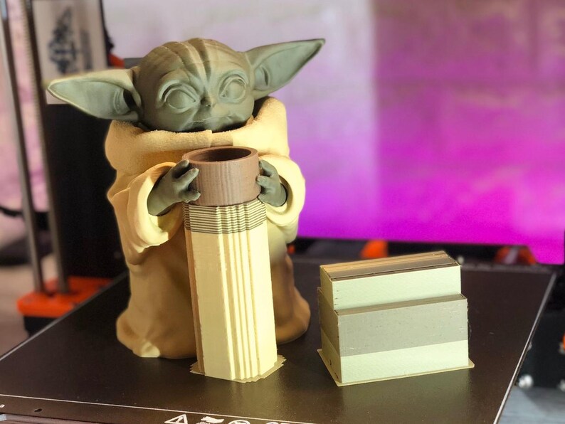 Pode incluir: Uma figura impressa em 3D de um personagem de uma popular franquia de fic&ccedil;&atilde;o cient&iacute;fica, segurando um objeto cil&iacute;ndrico. A figura &eacute; verde com orelhas grandes e est&aacute; vestindo uma t&uacute;nica marrom. Uma pilha de objetos retangulares est&aacute; ao lado da figura.
