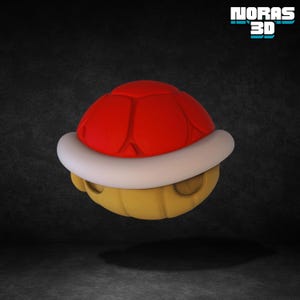 May include: A 3D rendered red Koopa Troopa shell with a white trim and a yellow underside. The shell is a recognizable element from the Super Mario video game series. The image includes the text "NORAS 3D".