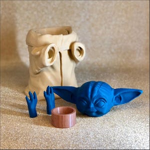 Pode incluir: Uma cole&ccedil;&atilde;o de itens com tema Star Wars impressos em 3D. Inclui uma cabe&ccedil;a azul de Grogu, duas m&atilde;os azuis, um anel de madeira e uma t&uacute;nica bege. Os itens est&atilde;o dispostos em uma superf&iacute;cie brilhante.