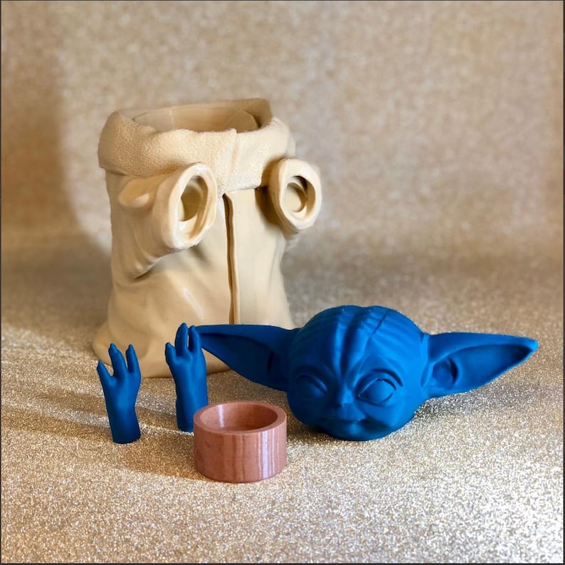 Pode incluir: Uma cole&ccedil;&atilde;o de itens com tema Star Wars impressos em 3D. Inclui uma cabe&ccedil;a azul de Grogu, duas m&atilde;os azuis, um anel de madeira e uma t&uacute;nica bege. Os itens est&atilde;o dispostos em uma superf&iacute;cie brilhante.