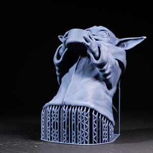 Pode incluir: Figura impressa em 3D azul claro de um personagem de uma franquia de fic&ccedil;&atilde;o cient&iacute;fica, segurando uma x&iacute;cara. A figura &eacute; suportada por uma base de treli&ccedil;a. A figura tem orelhas grandes e est&aacute; vestindo uma t&uacute;nica. O fundo &eacute; preto.