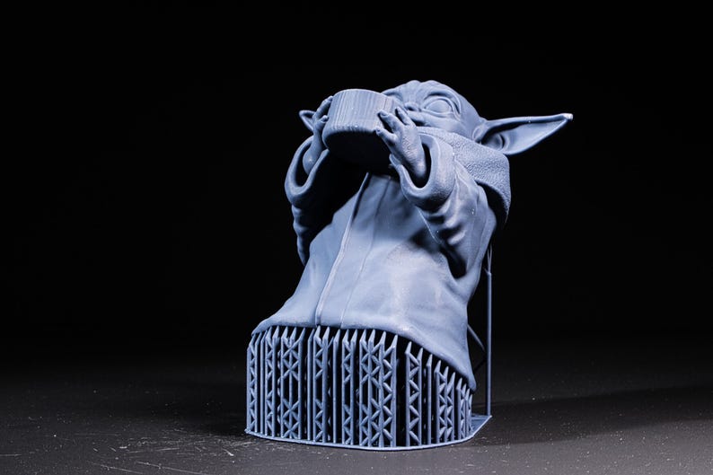 Pode incluir: Figura impressa em 3D azul claro de um personagem de uma franquia de fic&ccedil;&atilde;o cient&iacute;fica, segurando uma x&iacute;cara. A figura &eacute; suportada por uma base de treli&ccedil;a. A figura tem orelhas grandes e est&aacute; vestindo uma t&uacute;nica. O fundo &eacute; preto.