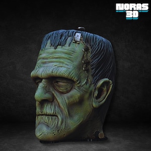 May include: A green and black Frankenstein head sculpture with visible stitches and textured skin. The sculpture has dark hair and a stern expression. The text "NORAS 3D" is displayed in blue and white.