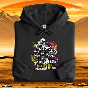 May include: Black hooded sweatshirt with a motorcycle graphic. The graphic features a rider on a motorcycle with the text "I'VE GOT 99 PROBLEMS BUT MY BIKE SOLVES MOST OF THEM!" in white and yellow. The background has pink and yellow paint splatters.